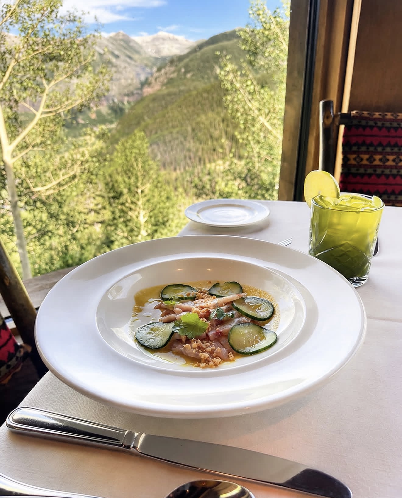 A cucumber crudo course plated at Allred's, with a cocktail and mountain views beyond the window