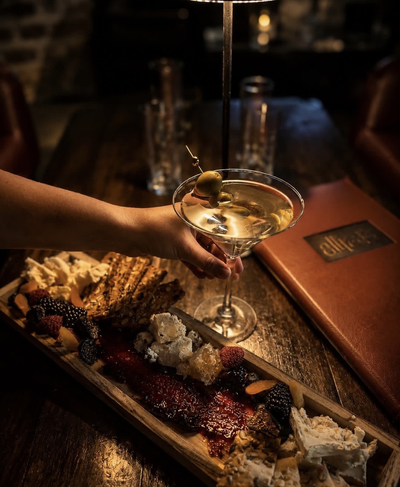 A martini and charcuterie board at Allred's, the leather menu embossed with the Allred's wordmark on the table beside it